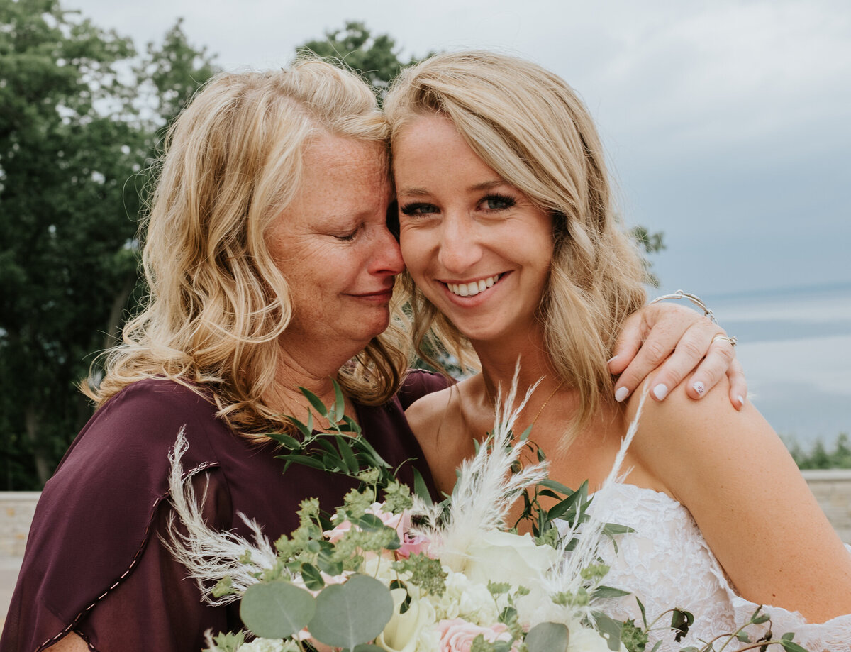 A mother holds her daughter, a bride, around the shoulder. They are both crying. The bride is also smiling.