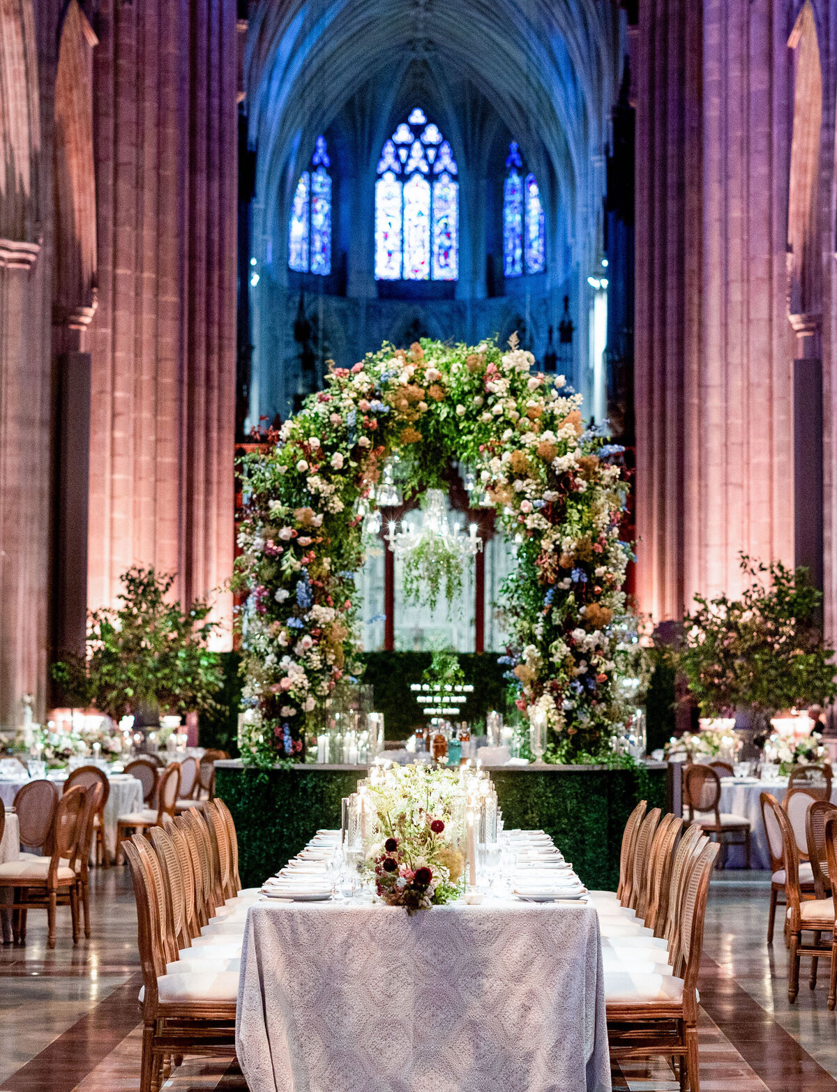 The photo was taken at Washington National Cathedral in Washington D.C. by photographer Twah Dougherty. The setting showcases a beautifully decorated wedding reception in a grand cathedral. The long dining table is adorned with white tablecloths, elegant tableware, and candles. A stunning floral arch made of colorful flowers stands in the background, with intricate stained glass windows above. You can add Current Films as Videographers to this information to capture the beautiful moments of the wedding reception.