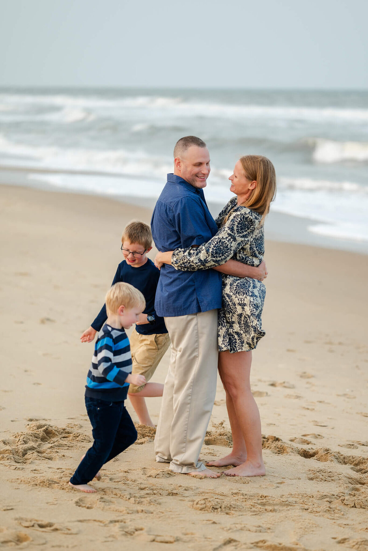 Mom and Dad hug each other in front of the waves on the beach while their two young sons run circles around them.