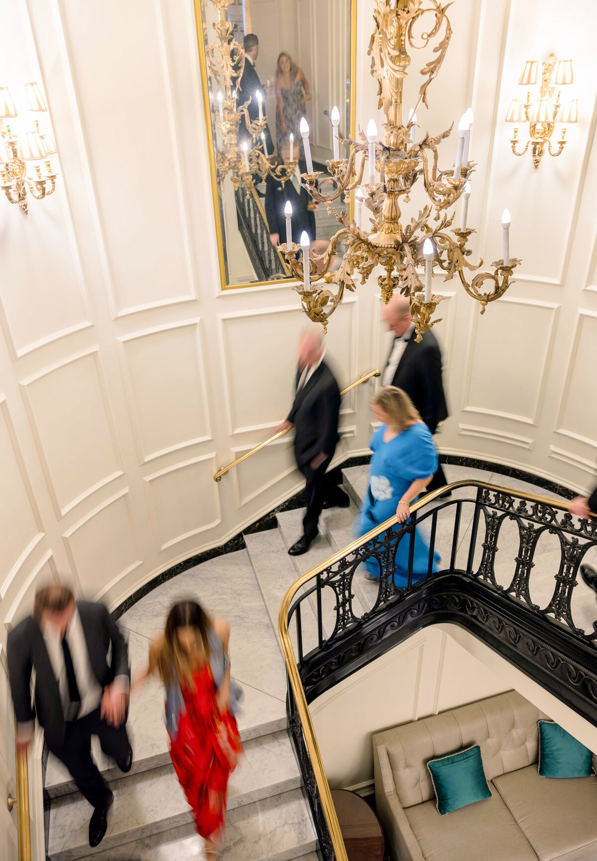 A group of formally dressed people descend an ornate spiral staircase beside a large chandelier. The white-paneled walls and a mirror reflect some of the individuals. The focus is soft, capturing a sense of movement.
