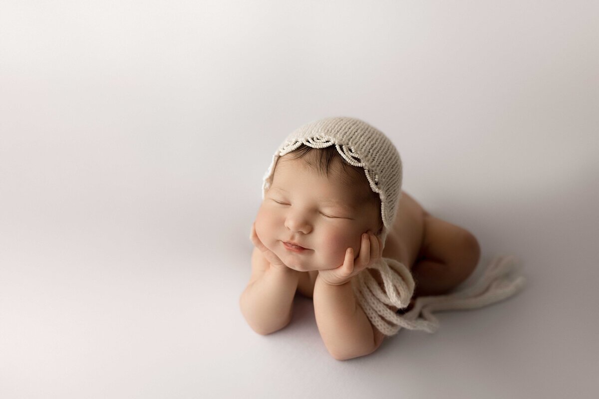 Best Newborn Photographer in London, ON. Baby in crochet bonnet is sitting in froggy pose with her hands resting under her cheek.
