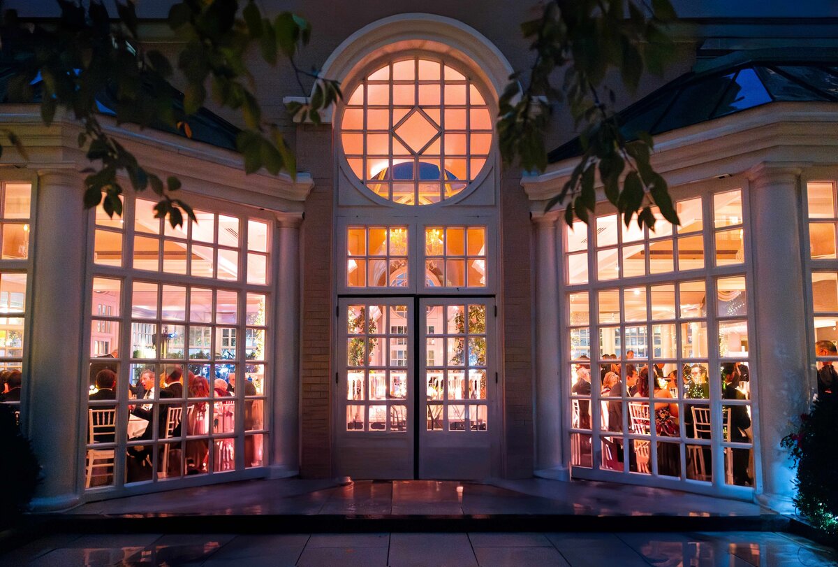 Elegant event venue with large arched windows glowing with warm light. Inside, guests are gathered at tables, suggesting a festive occasion, possibly a wedding or banquet. Trees frame the scene, adding a touch of nature to the ambiance.