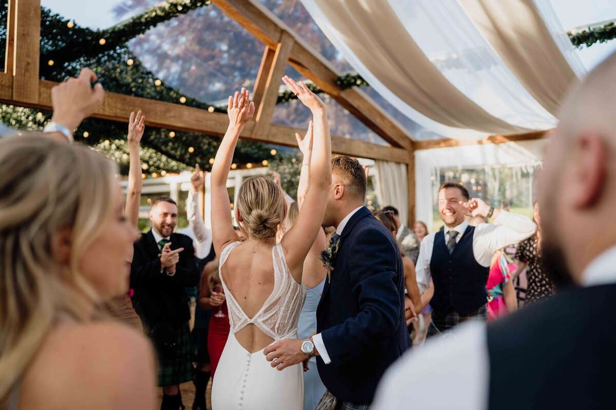 A joyful wedding reception scene photographed by an Aberdeen wedding photographer at House of Elrick, with people dancing under a canopy adorned with greenery and string lights. The bride, in a white dress, is dancing with a man in a suit while guests cheer and laugh in the background.