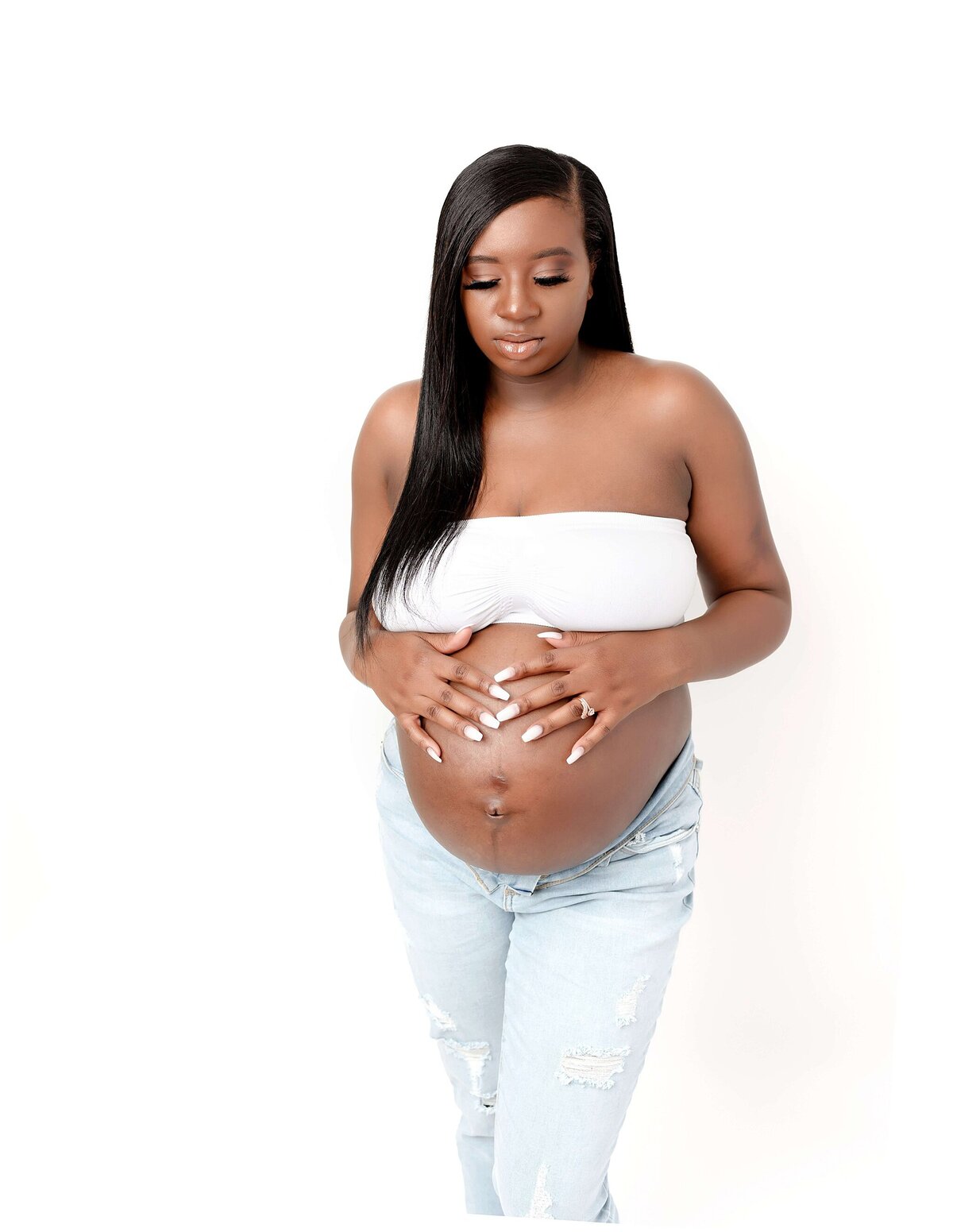 Pregnant woman standing with her hands gently resting on her rounded belly, showcasing the bond between mother and unborn child. She wears jeans and a tube top.