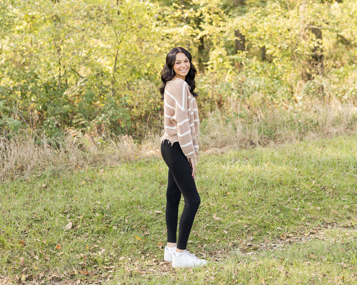 High school female, standing in grass with a tree line behind her, turned to the side, smiling at the camera, with long black hair, wearing black leggings, white tennis shoes, and tan and white striped long sleeve sheet.