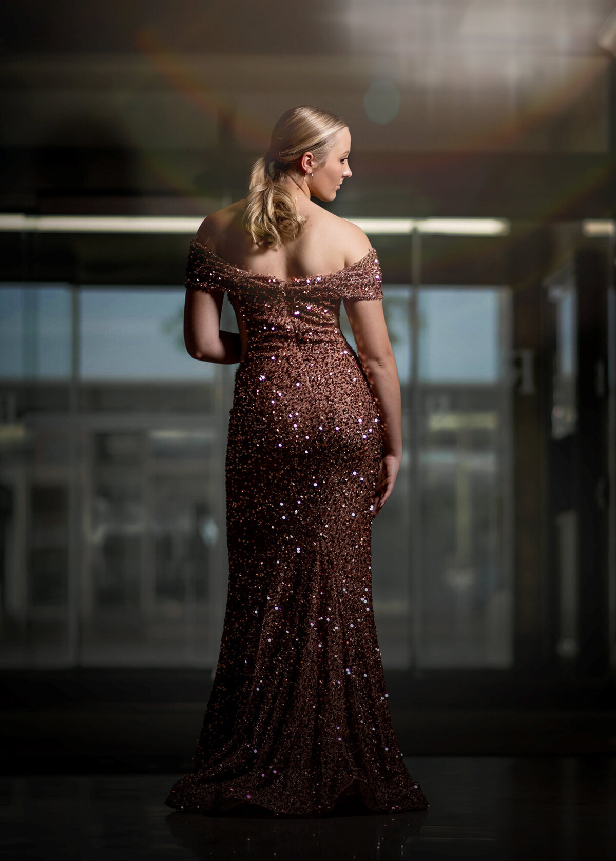 A blonde high school senior stands in a tall glass window in an elegant gown