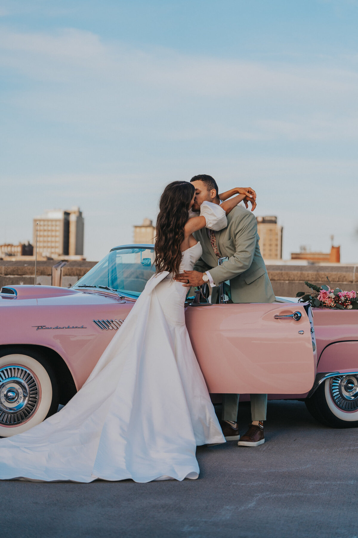 A woman leans over the driver door of a pink convertible and kisses her husband