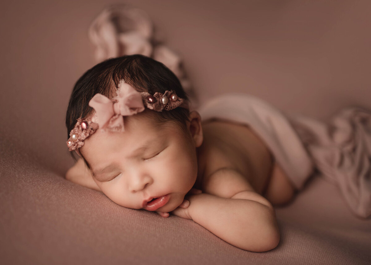 Baby sleeping on belly with hands under head on rose gold knit. Baby wearing headband with blow and pearl details.