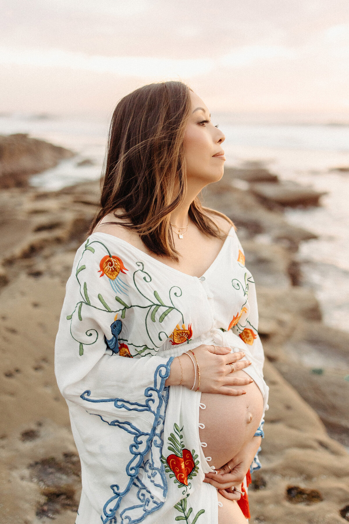 Pregnant mother in a floral embroidered gown looking out at the ocean during a maternity photoshoot in La Jolla. Captured by San Diego Family Photographer Yuka Schad.