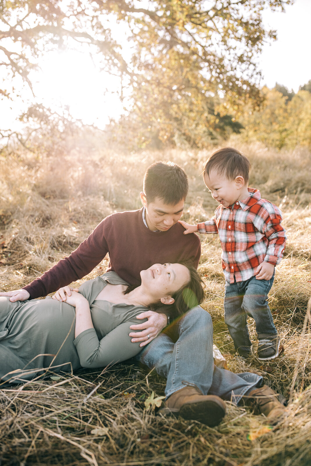 Family maternity photo session with mom laying her head on her husband's lap looking at each other, their son standing to the side looking at mom and dad