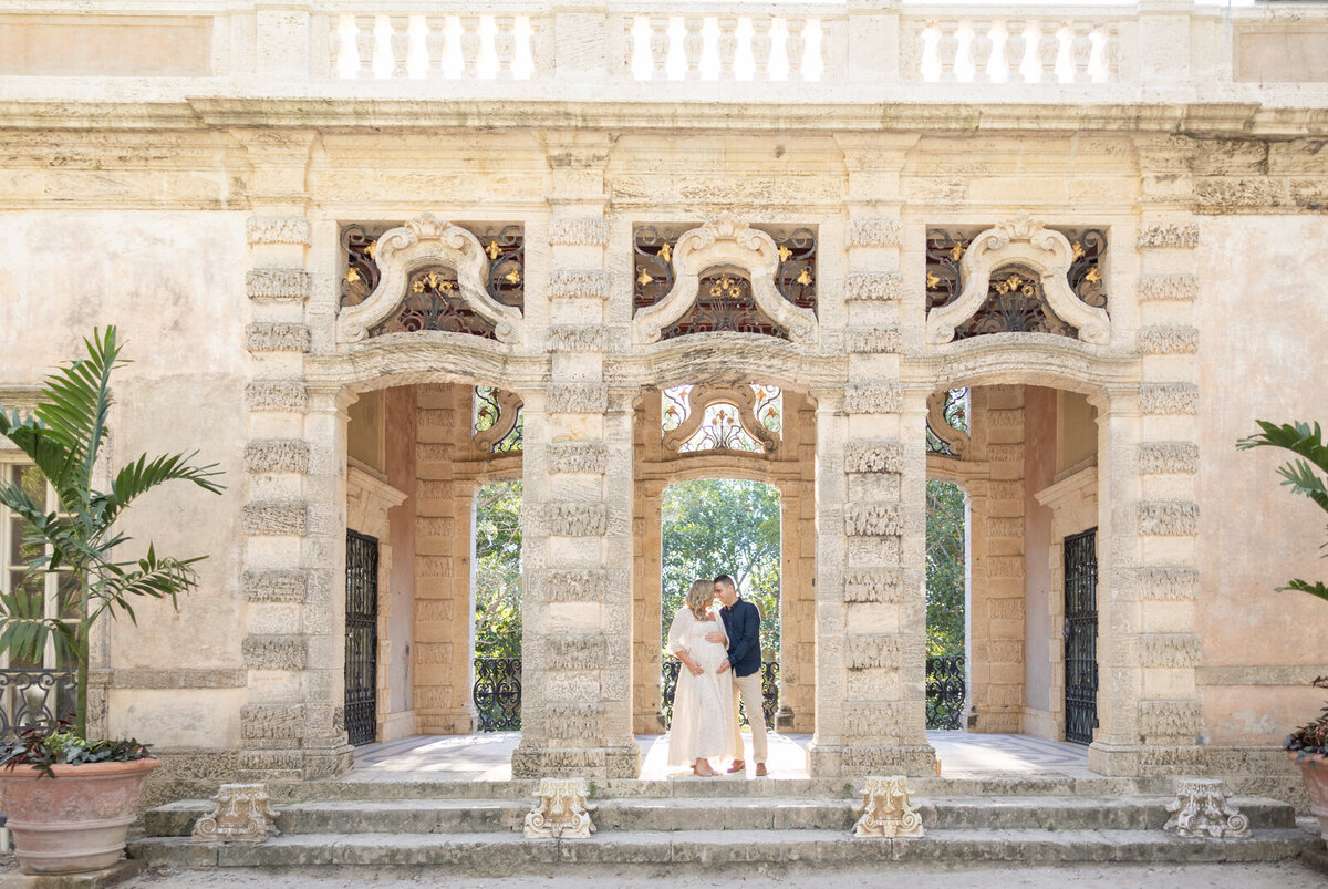 A beautiful expectant mother in a white dress, standing on a grand balcony with a scenic backdrop.