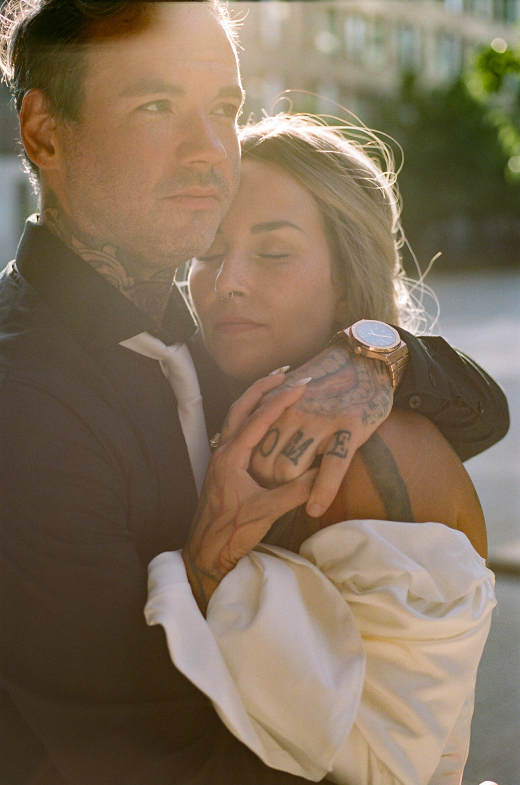 Man and woman embrace outdoors, captured beautifully by an Oshawa engagement photographer. The man displays tattoos on his neck and hand, pairing a black shirt with a white tie. The woman, eyes closed in serenity, dons a white top and sports a watch on her wrist.