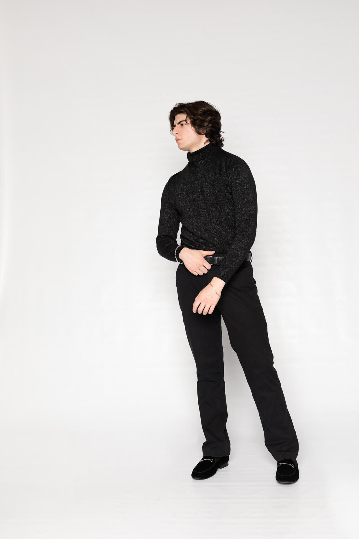 A person with wavy hair stands against a plain white background, wearing a dark turtleneck sweater and dark pants. They are looking to the side with one hand placed on their hip.