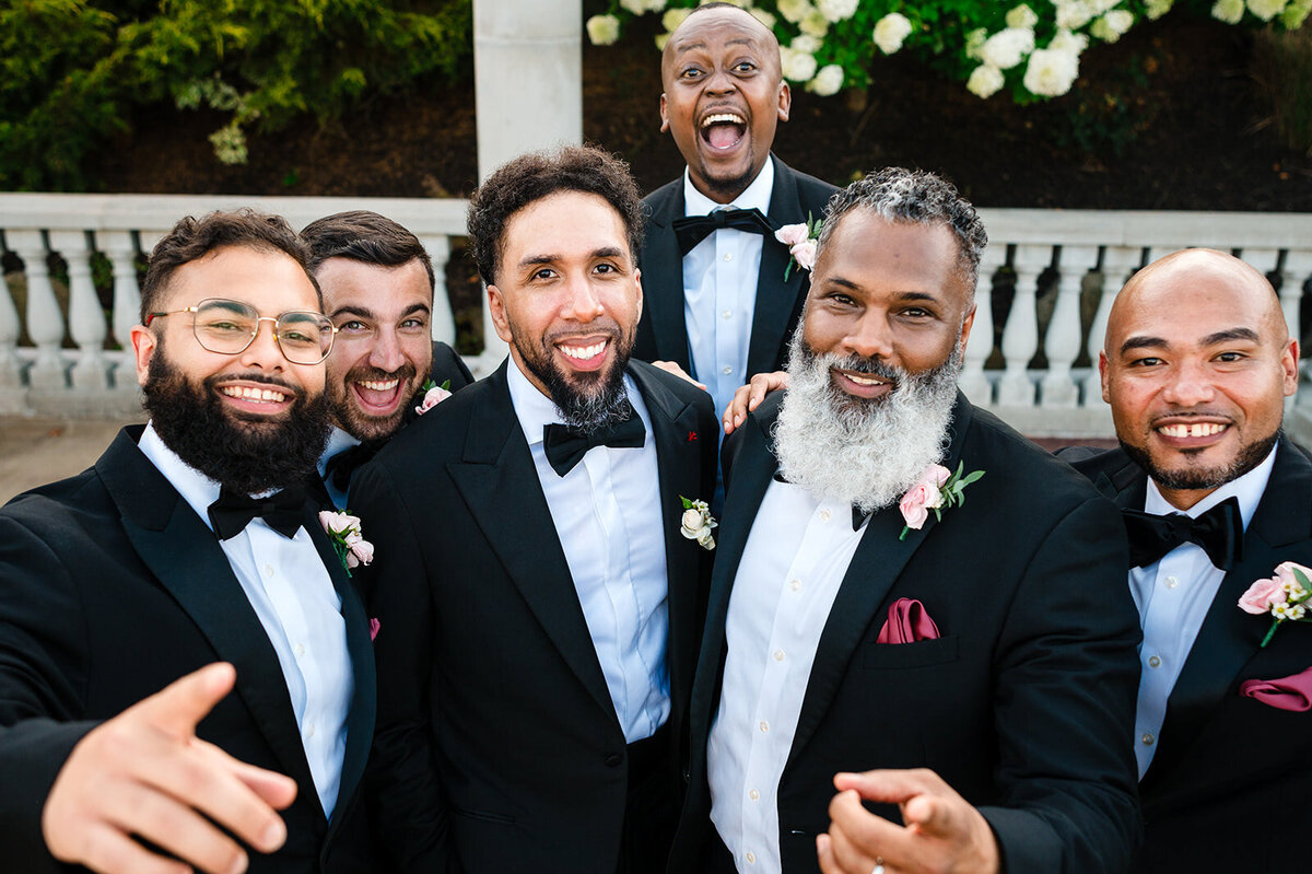 Groom and groomsmen in black tuxedos with burgundy pocket squares taking a cheerful selfie