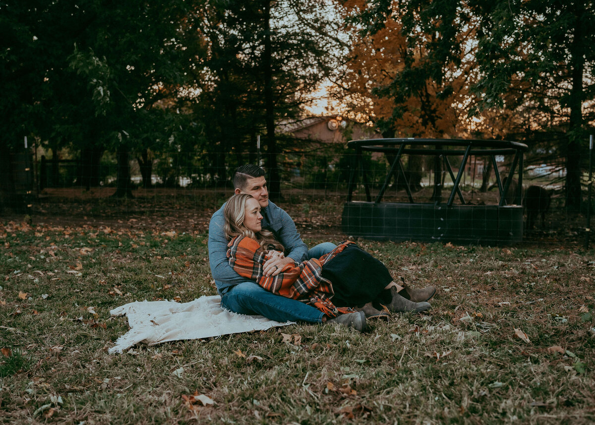 A farmer holds his wife on a blanket as they cozy up with their gorgeous family farm in the backdrop during the beautiful autumn colors.