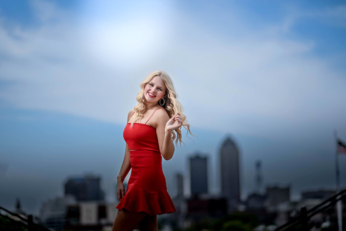 A high school senior in a red dress walks with city skyline twirling her hair