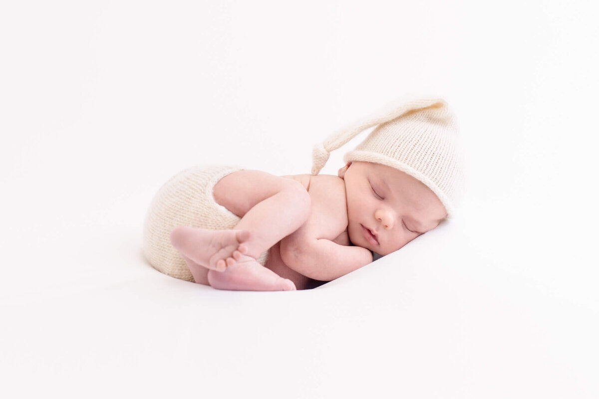 A newborn baby peacefully sleeps curled up on a white background, wearing a soft, knitted cream hat with a long tail and matching diaper cover. The scene conveys a sense of innocence and tranquility.