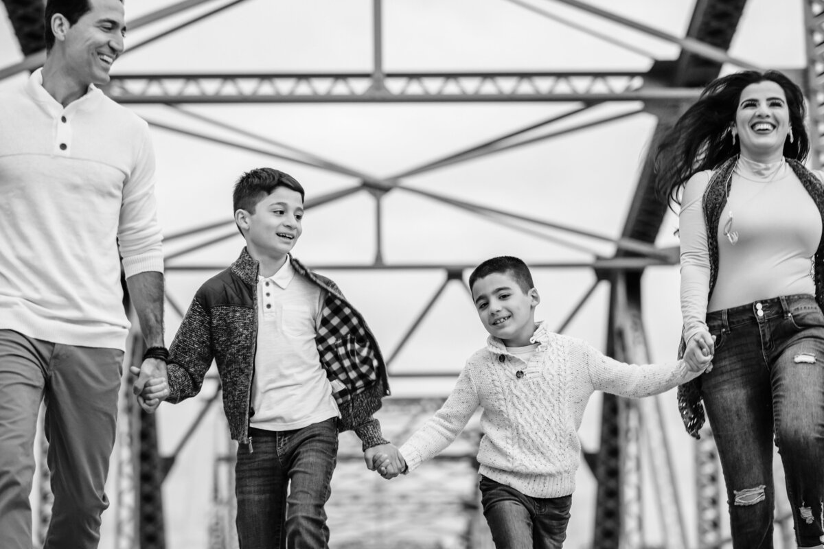 A black-and-white photo of a family of four walking hand in hand on a metal bridge. The parents walk on the outside, with the two young boys in the center, all smiling as they walk towards the camera.