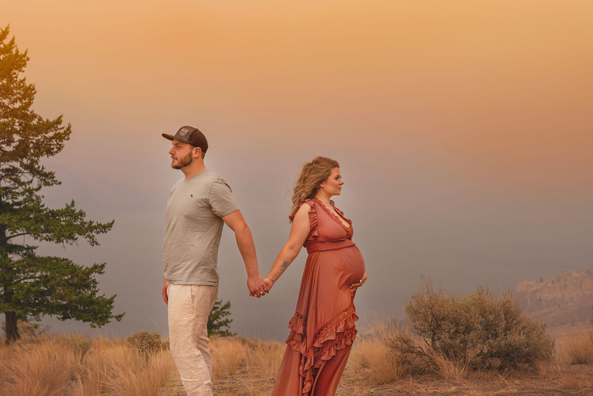 A man and pregnant woman holds hands while looking in opposite directions. Behind them is a smokey sky sunset in Tobiano, Kamloops. Photo by Brittany Danielle