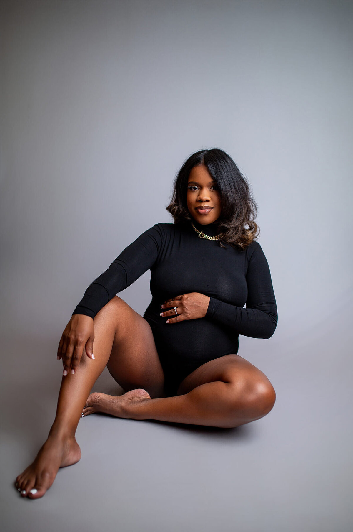 Black woman in black body maternity suit with hand on belly and other hand resting on knee.