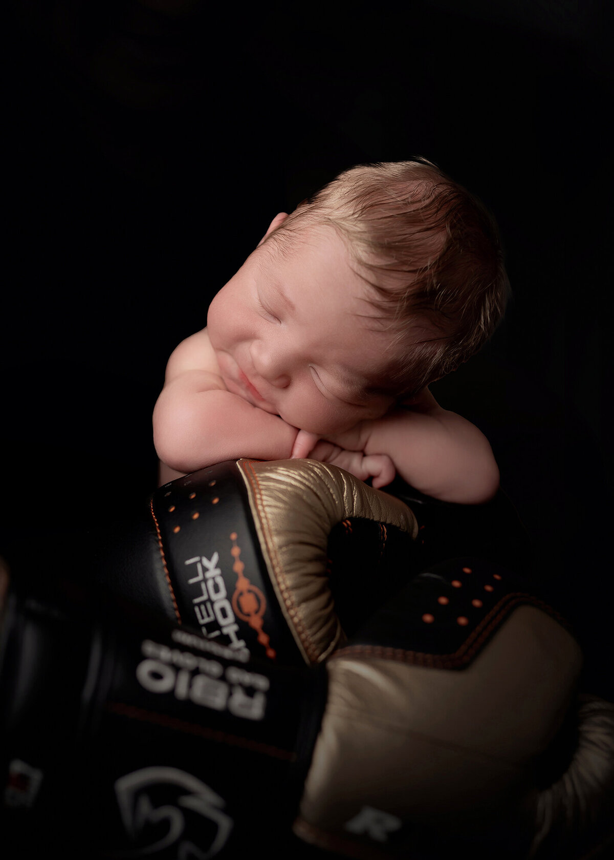 A newborn baby sleeps atop black boxing gloves in the studio of a Pella Newborn Photographer