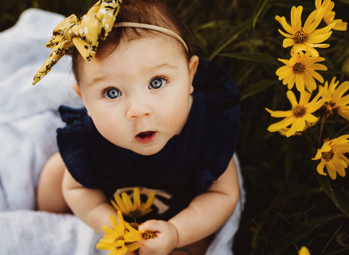 A baby girl is looking up at the camera while holding wildflowers.