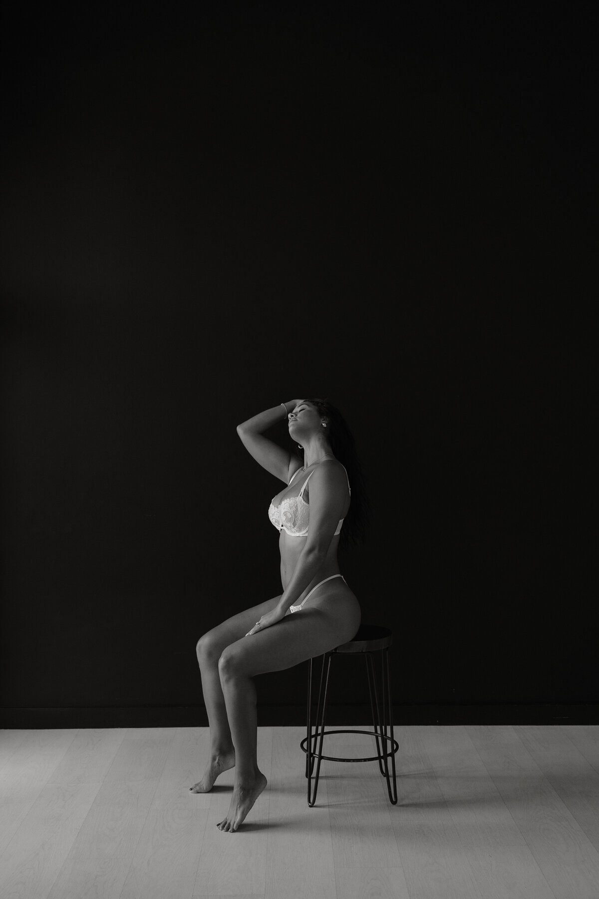 Black and white photo of woman in white lingerie sitting on a stool in front of black wall
