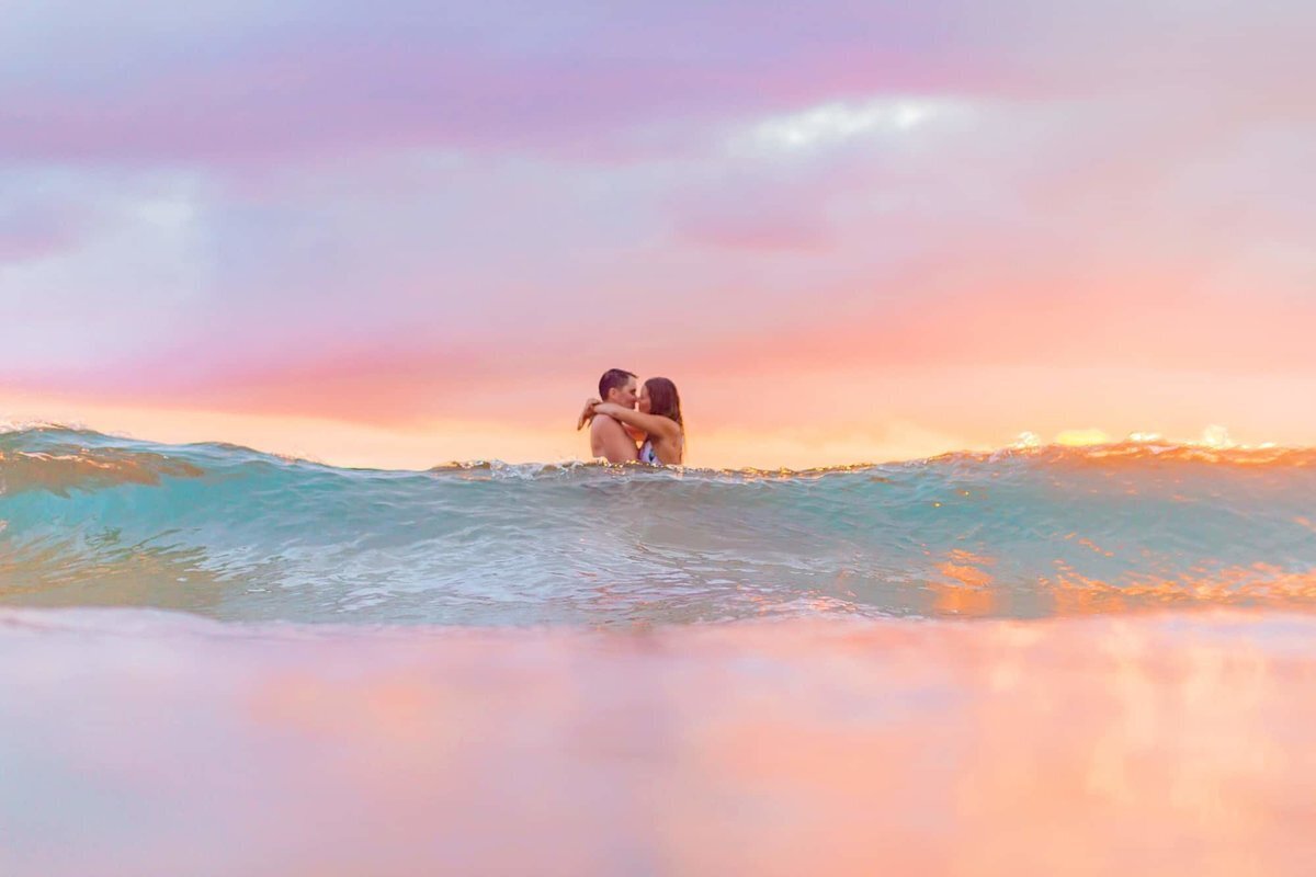 Romantic and colorful water engagement session with Love + Water, wave washes over the couple who are kissing in the shorebreak