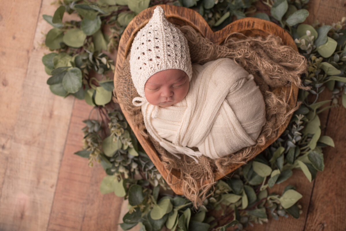 Baby boy wrapped in white in heart bowl with greenery around him