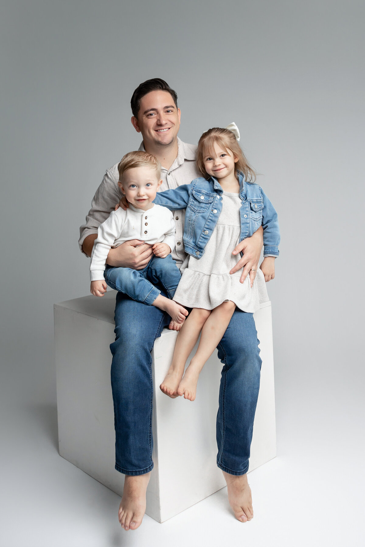 Father sitting on stool with his daughter on his lap, both smiling in simple studio setting. Photographed by Yucaipa family photographer Kaitlyn Dawn Photography.