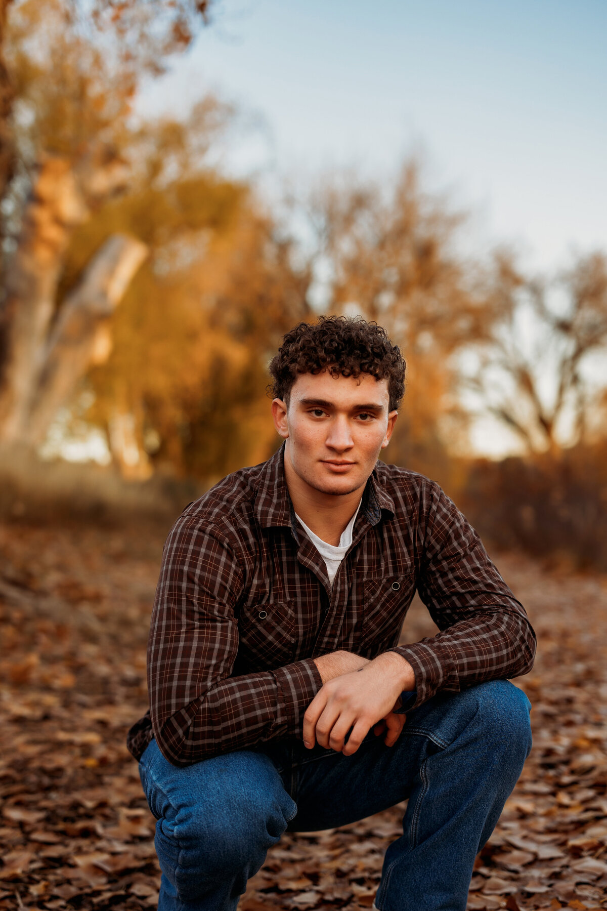 Senior boy squatting on a path covered in leaves in jeans and plaid button up for high school senior photos
