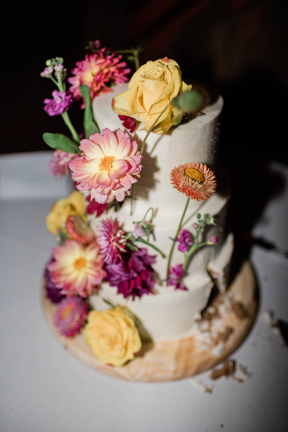 A three-tiered wedding cake adorned with fresh yellow, pink, and purple flowers, with buttercream frosting.