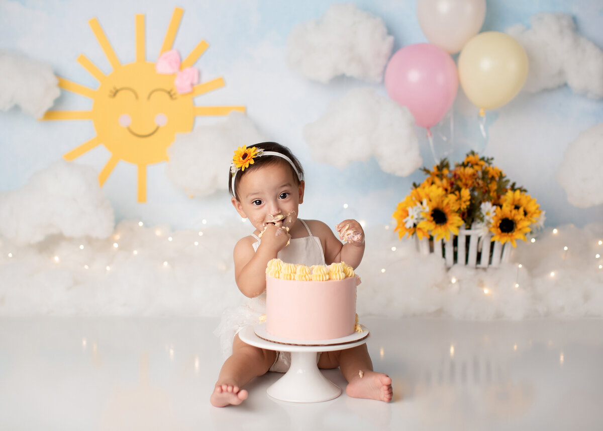 Sunshine themed cake smash at West Palm Beach photographer studio. Baby girl is sitting behind blush and yellow cake taking a bit of icing and smiling at the camera. In the background, there is a cute cartoon sunshine, faux clouds, and balloons.