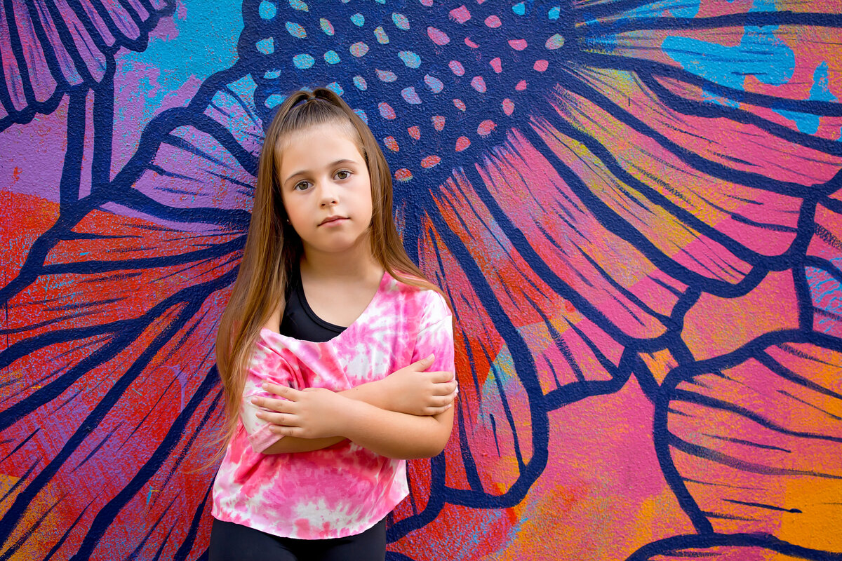 Young girl wearing pink tie dye shirt is standing in frown of floral mural in Downtown Riverside