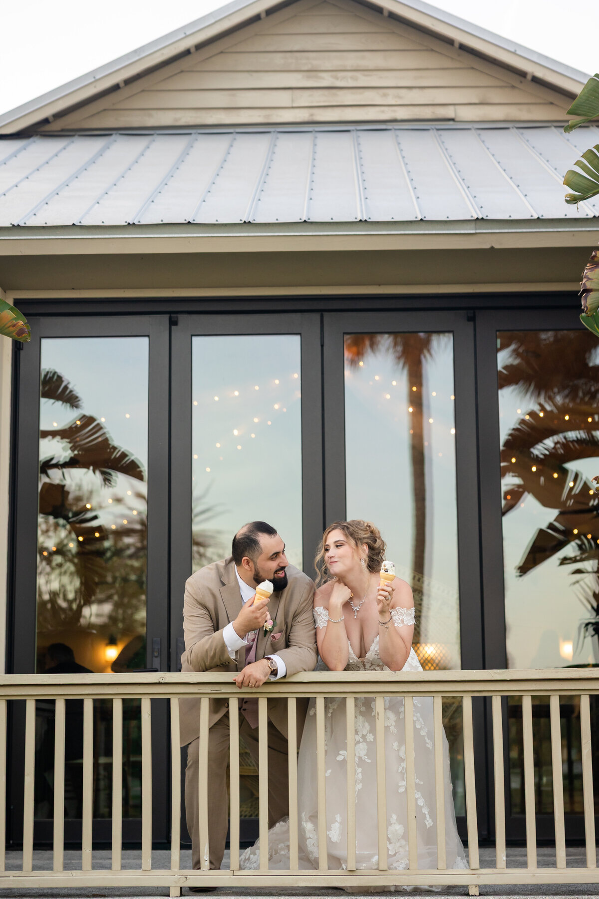 A bride and groom share a sweet, playful moment while enjoying ice cream cones on the balcony of their Jacksonville wedding venue. Reflections of palm trees and twinkle lights glow in the glass windows behind them, creating a romantic coastal vibe. The bride, in a lace off-the-shoulder gown, leans over the railing beside her groom, who wears a tan suit. Captured during a relaxed reception by a Jacksonville wedding photographer focused on authentic, candid wedding moments.