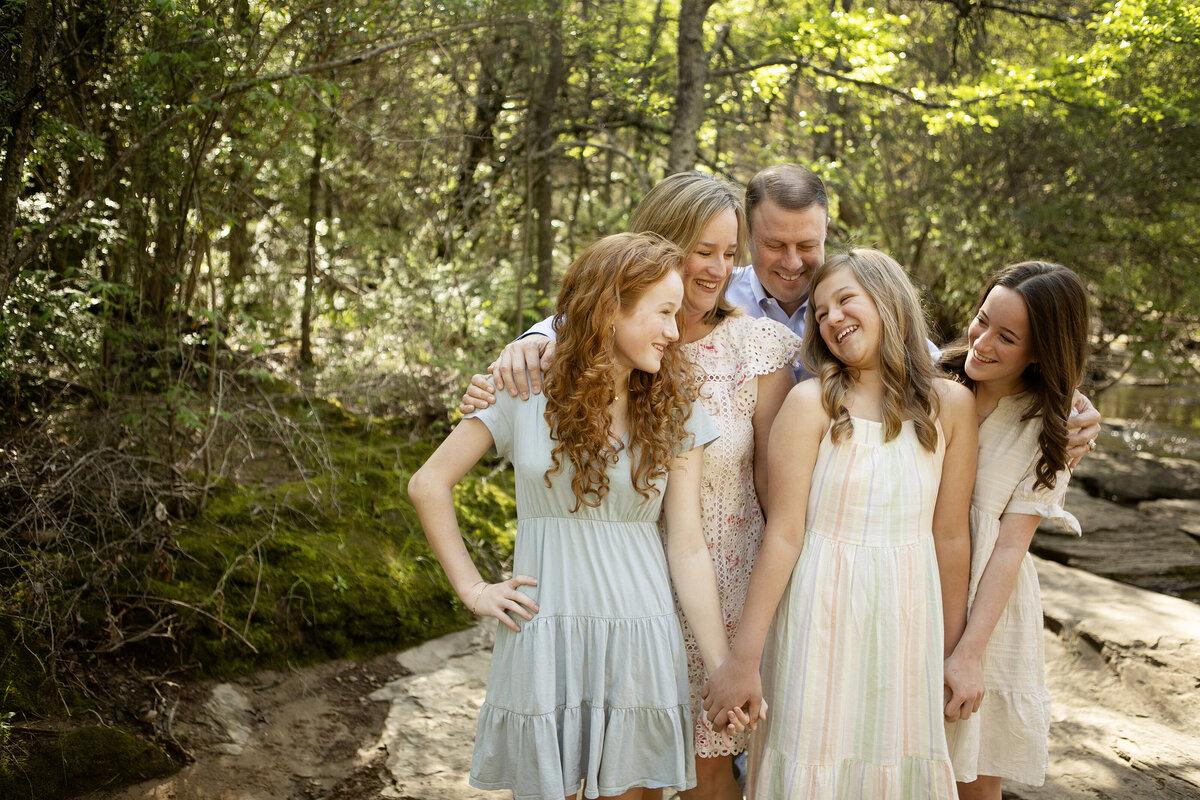 Three sisters standing together on the creek’s edge in Flower Mound, smiling as they pose for a photo