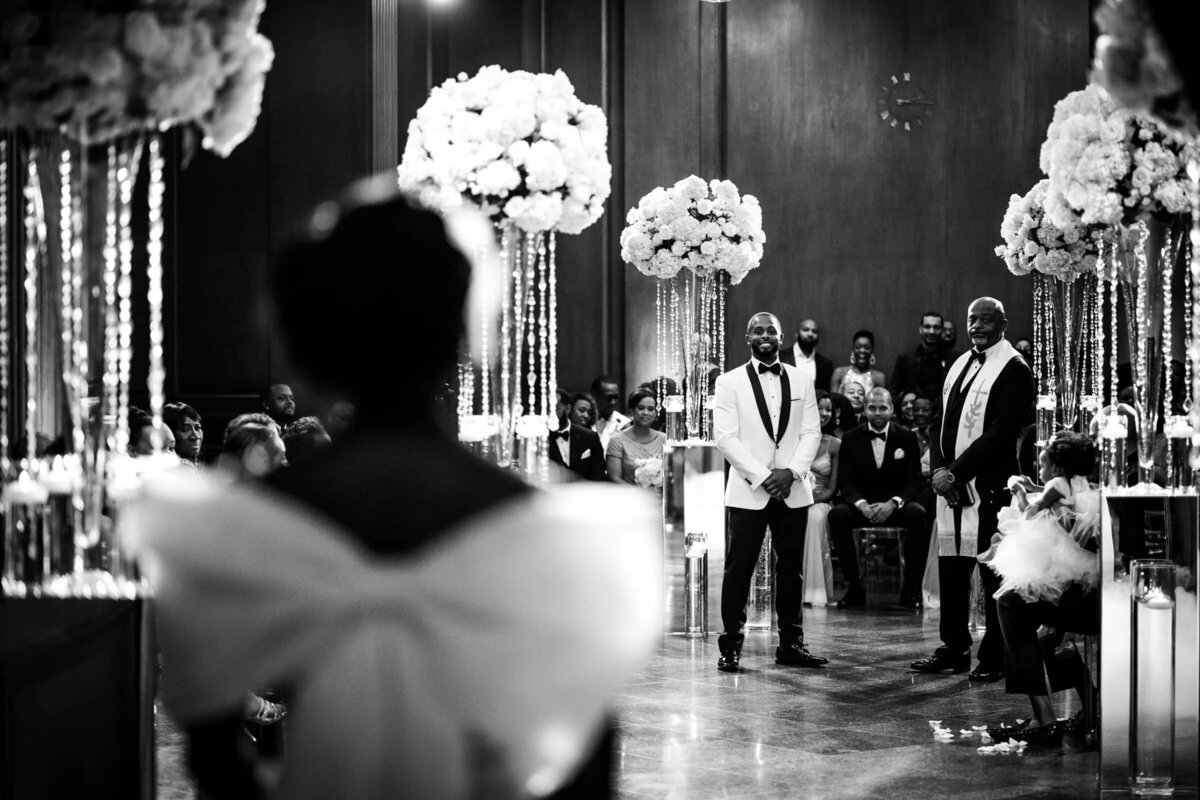 Bride in a white dress seen from the back, looking towards the groom and best man standing at the altar
