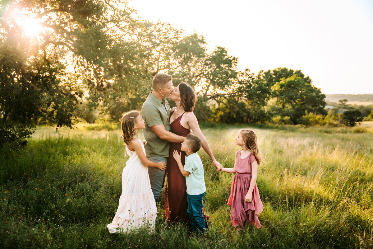 Family Photographer, mom and dad embrace as their kids admire them in a grassy field