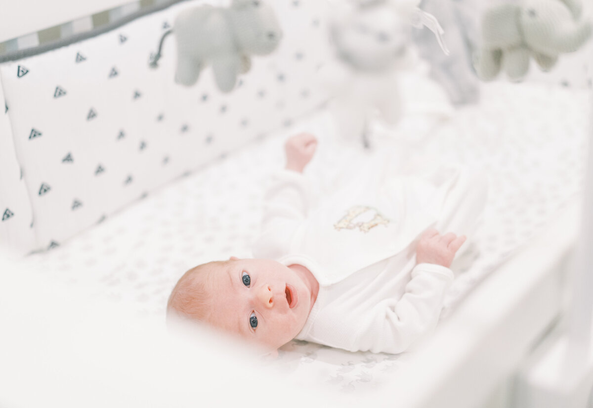 Portrait of a baby wearing a white onesie and bib lying awake in a white and gray bassinet while looking at a mobile up above