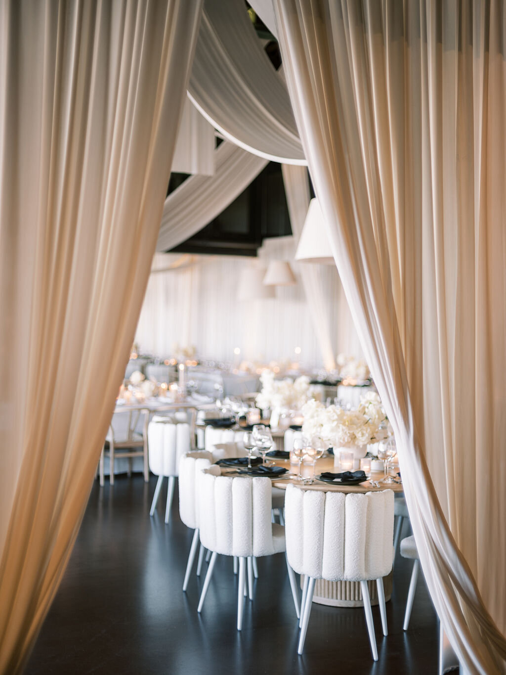 Flowing white drapery frames a luxurious wedding reception with modern white chairs and floral arrangements.