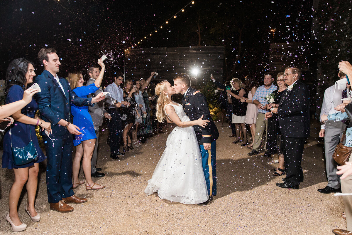 A bride and groom share a kiss during their exit after their wedding at BRIK Venue in Fort Worth, Texas. The bride is on the left and is wearing a sleeveless, intricate white dress. The groom is on the right and is wearing his military uniform. Guests stand on either side of them and shower them in confetti.