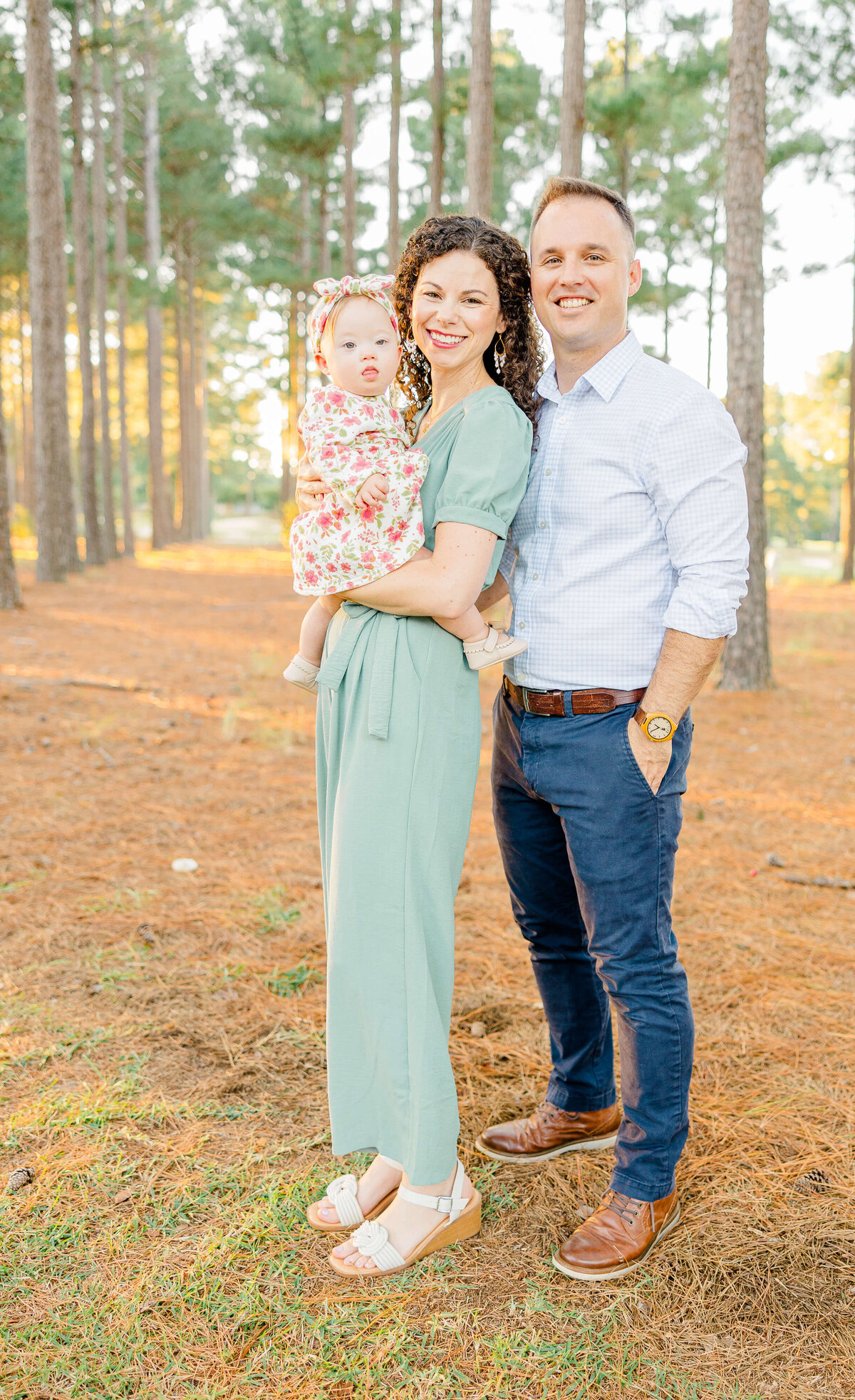 A woman in a green jumpsuit cradles a baby in floral attire, standing beside a man in light blue shirt and dark pants. They pose elegantly in a woodland area with sunlight filtering through—an ideal scene captured by a skilled Fayetteville NC family photographer.