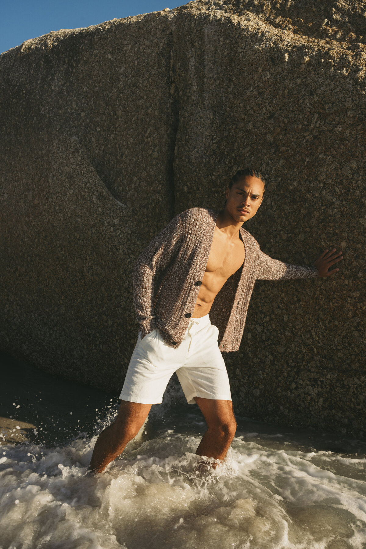Timothe standing in the ocean surf in Cape Town, wearing white shorts and an open knit cardigan, leaning against a large rock in bright sunlight.