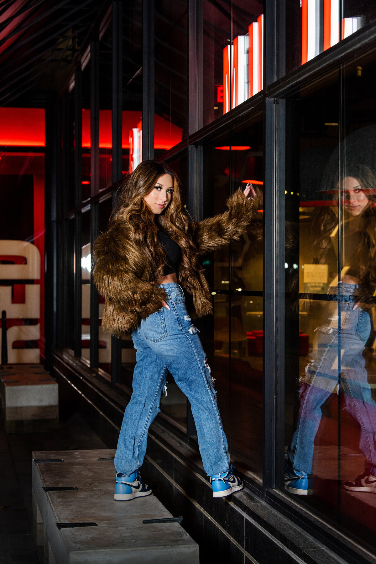 senior girl in jeans and fashion fur coat leaning against glass in urban setting
