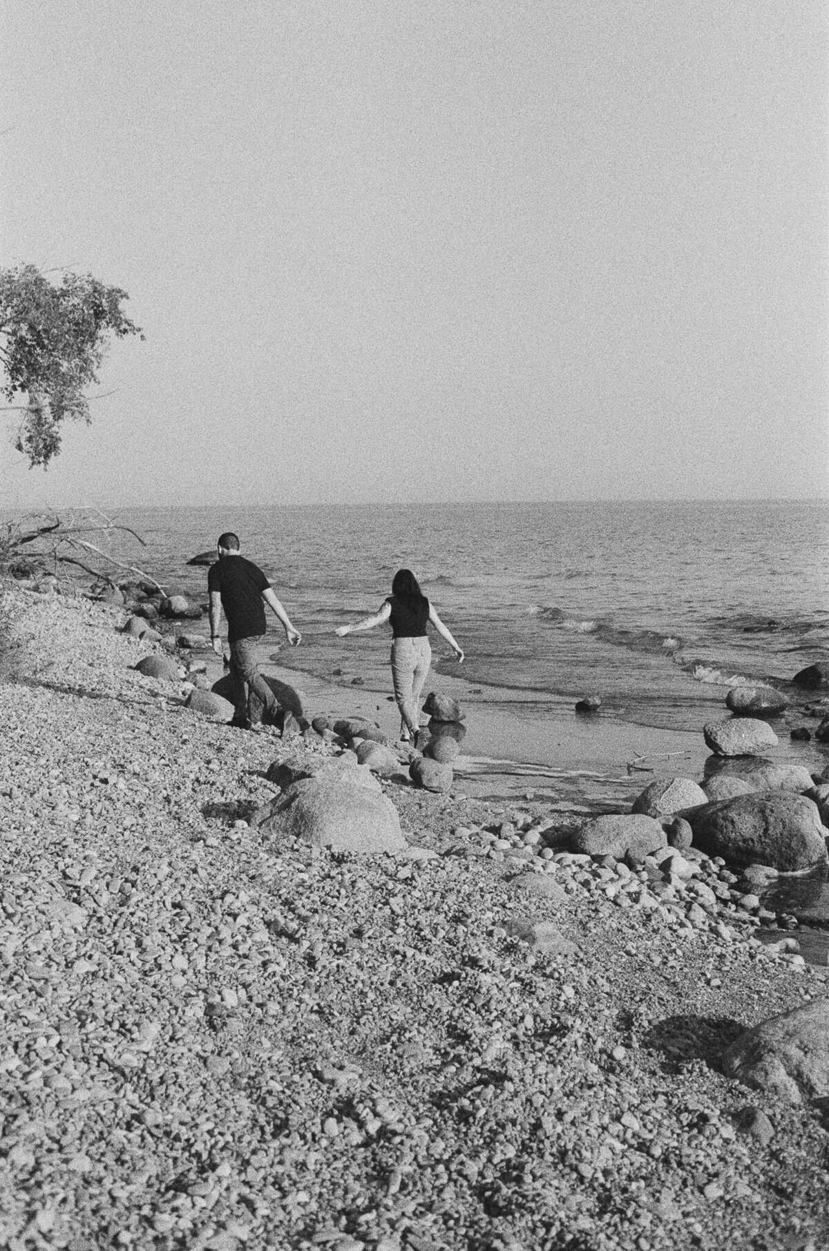 A couple walks hand in hand on a rocky beach shore in Oshawa, the sea serenely stretching behind them and a tree to the left. Captured in beautiful grayscale by Oshawa engagement photographer, Jennifer van Son, this image exudes timeless romance.