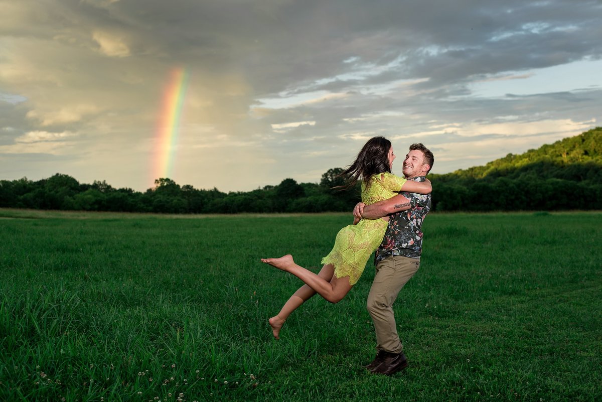 Bride wearing neon yellow lace dress being spun by her fiance in the middle of an open grass field at Drakewood Farm with sunsetting behind them and a rainbow showing