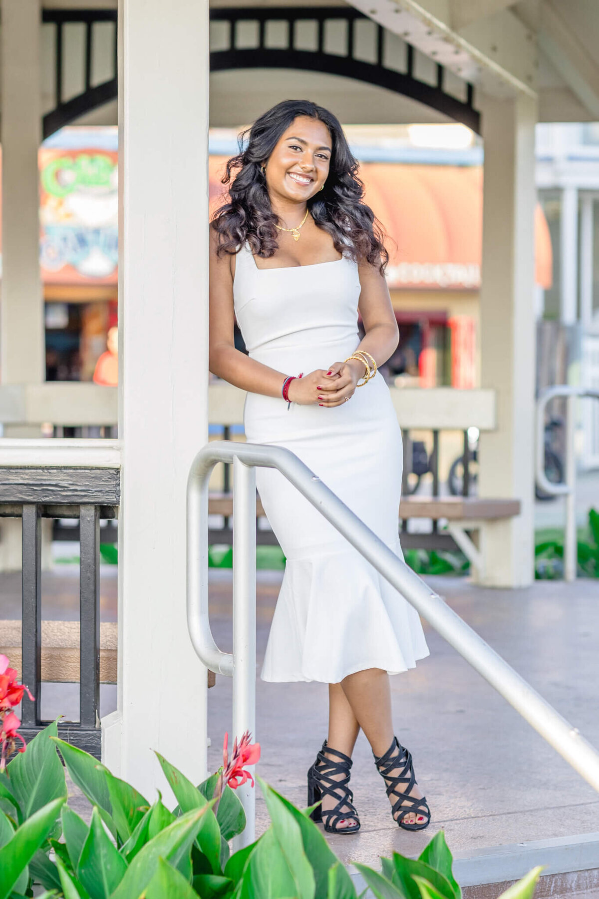 A woman in a white dress stands smiling under a gazebo. She is leaning against a wooden post with her hands clasped. There are red flowers and greenery around her, with a blurred background of storefronts.