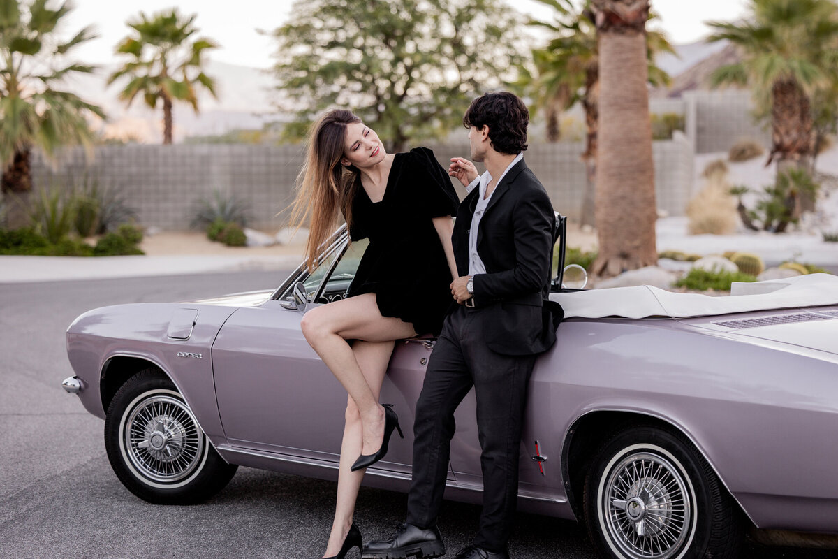 Laughing couple by vintage convertible during golden hour in the desert, surrounded by palm trees and desert tones.