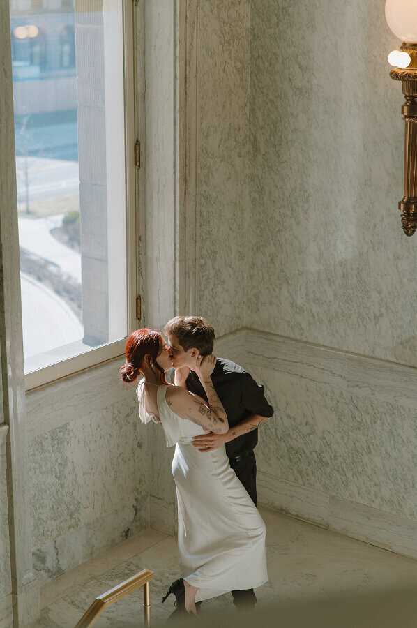 A man and woman kiss on their elopement day. The natural light from the window next to them is illuminating them.