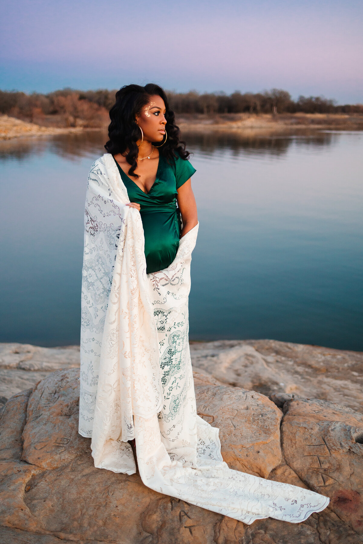 The beauty of maternity with this photograph by an Albuquerque maternity photo specialist. A pregnant woman stands gracefully on rocks beside a serene lake, dressed in a flowing green dress and a delicate white lace blanket.